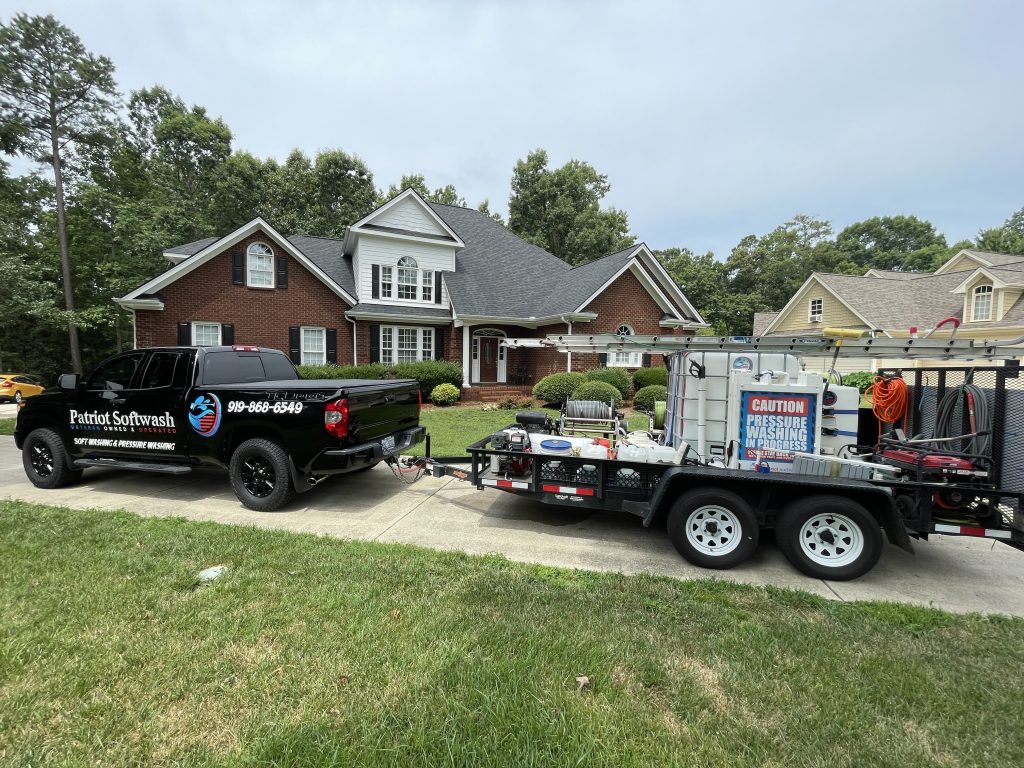 Patriot Softwash pressure washing truck and equipment trailer parked in front of a large brick home during a residential exterior cleaning project in Apex, NC.